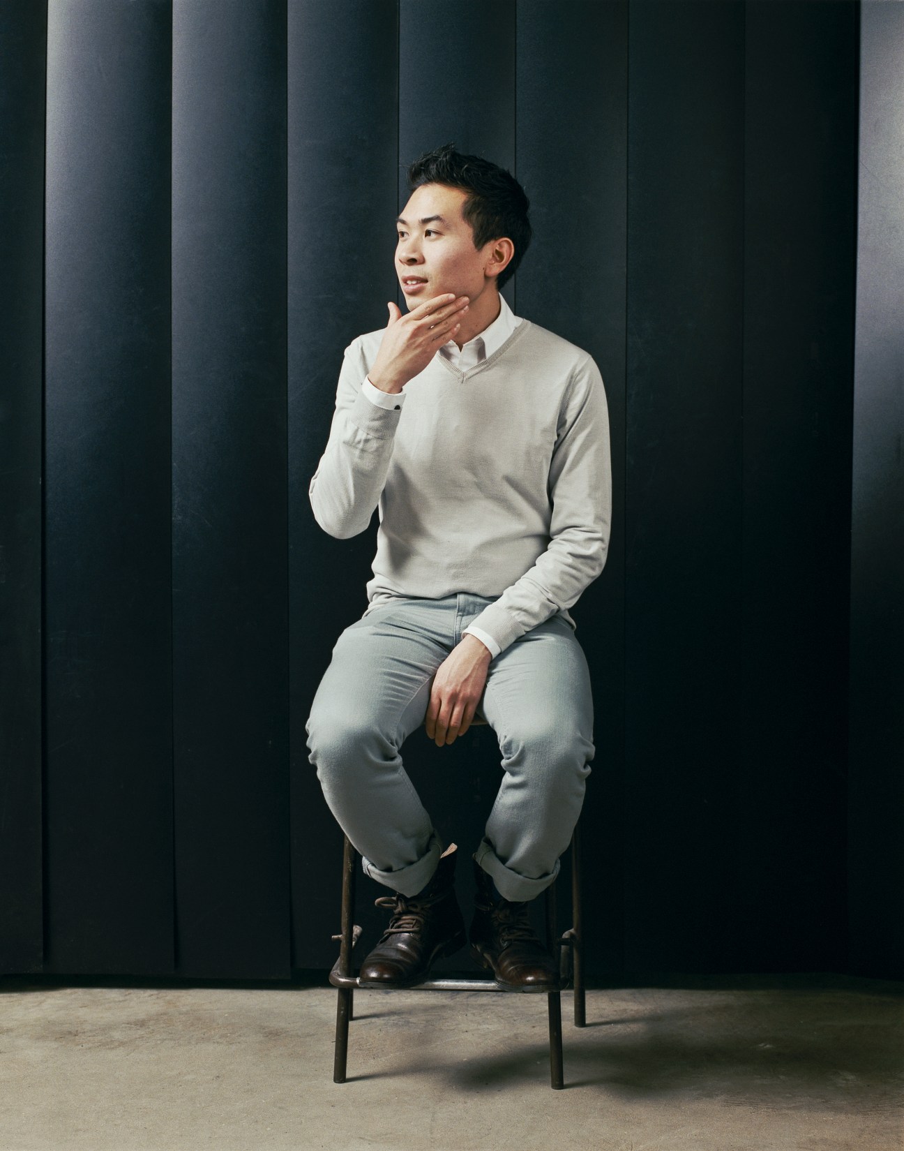 Vuong Tong, a male student in light-colored clothing, sits on a stool against a dark striped background.