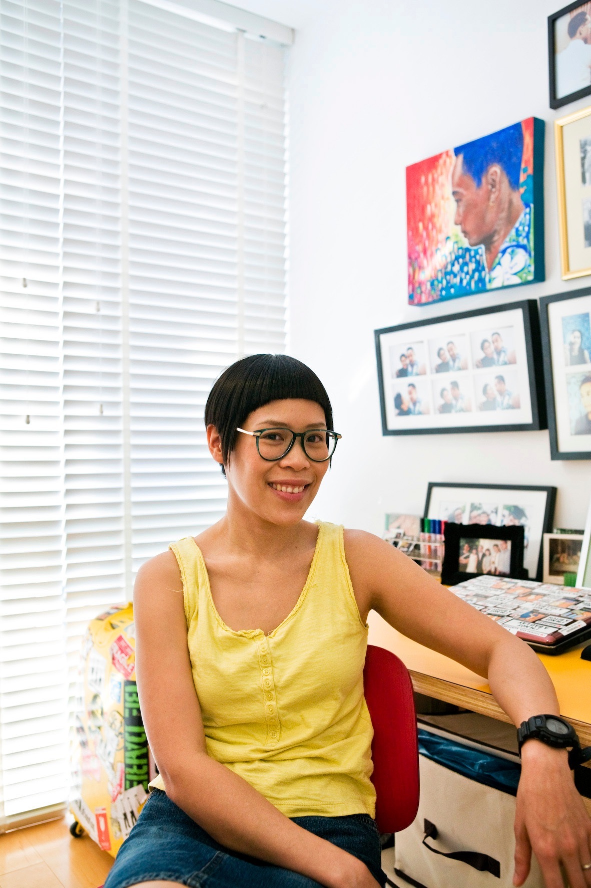 Pin Nakarabundit, an advertising professional, sitting in her office wearing a yellow tank top and glasses, smiling at the camera with framed photos on the wall behind her.