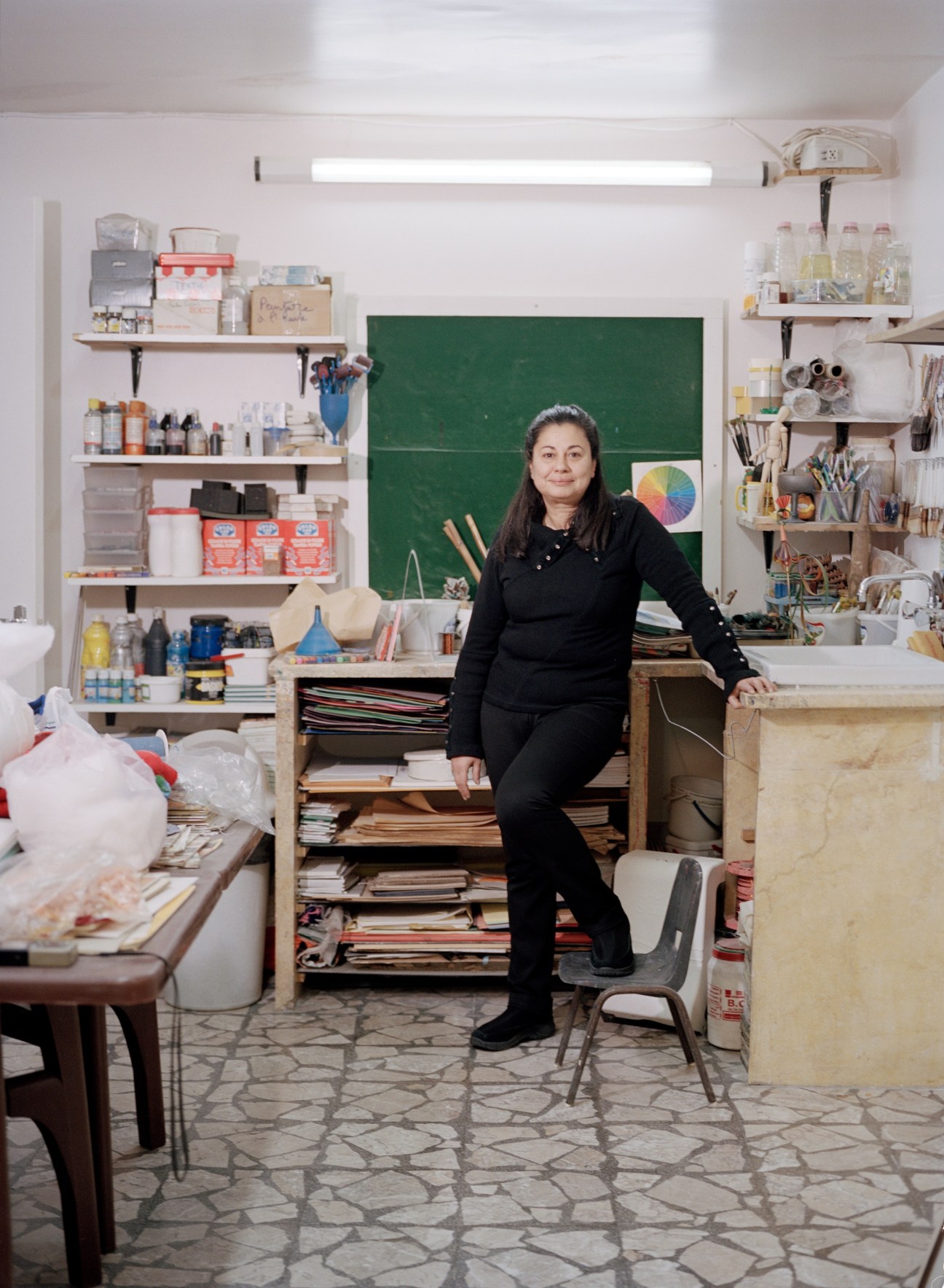 Art teacher Zena Sabbagh poses in her art studio surrounded by shelves of supplies and materials.