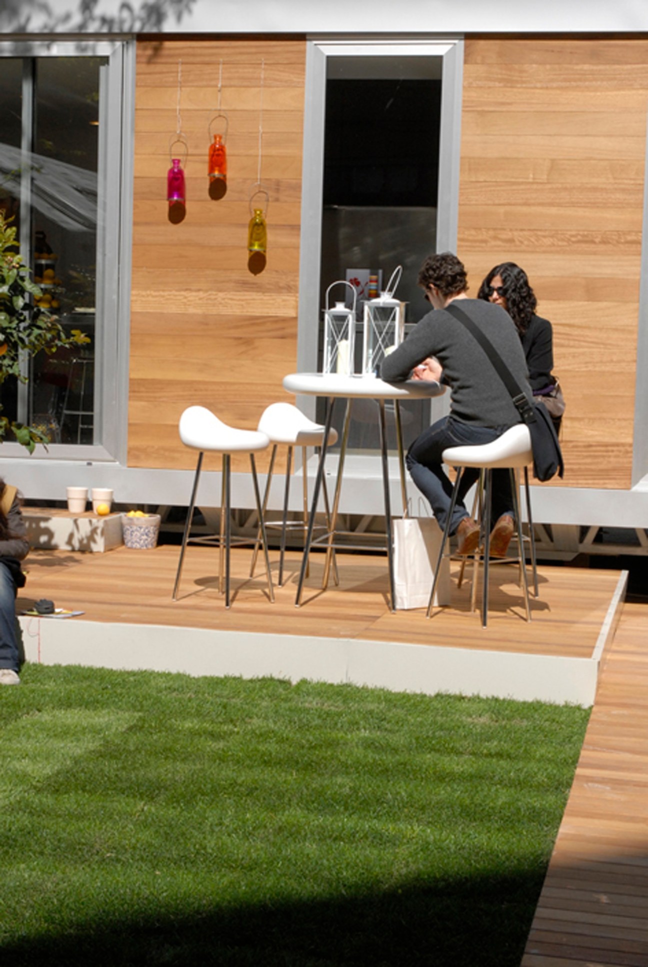 Two people sit at a small table with white chairs on a wooden terrace, with colorful hanging lights and wooden cladding visible behind them.