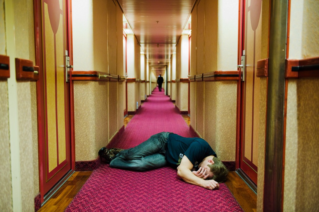 A person lying asleep in a ship corridor with red carpeting and cream-colored walls with red trim.