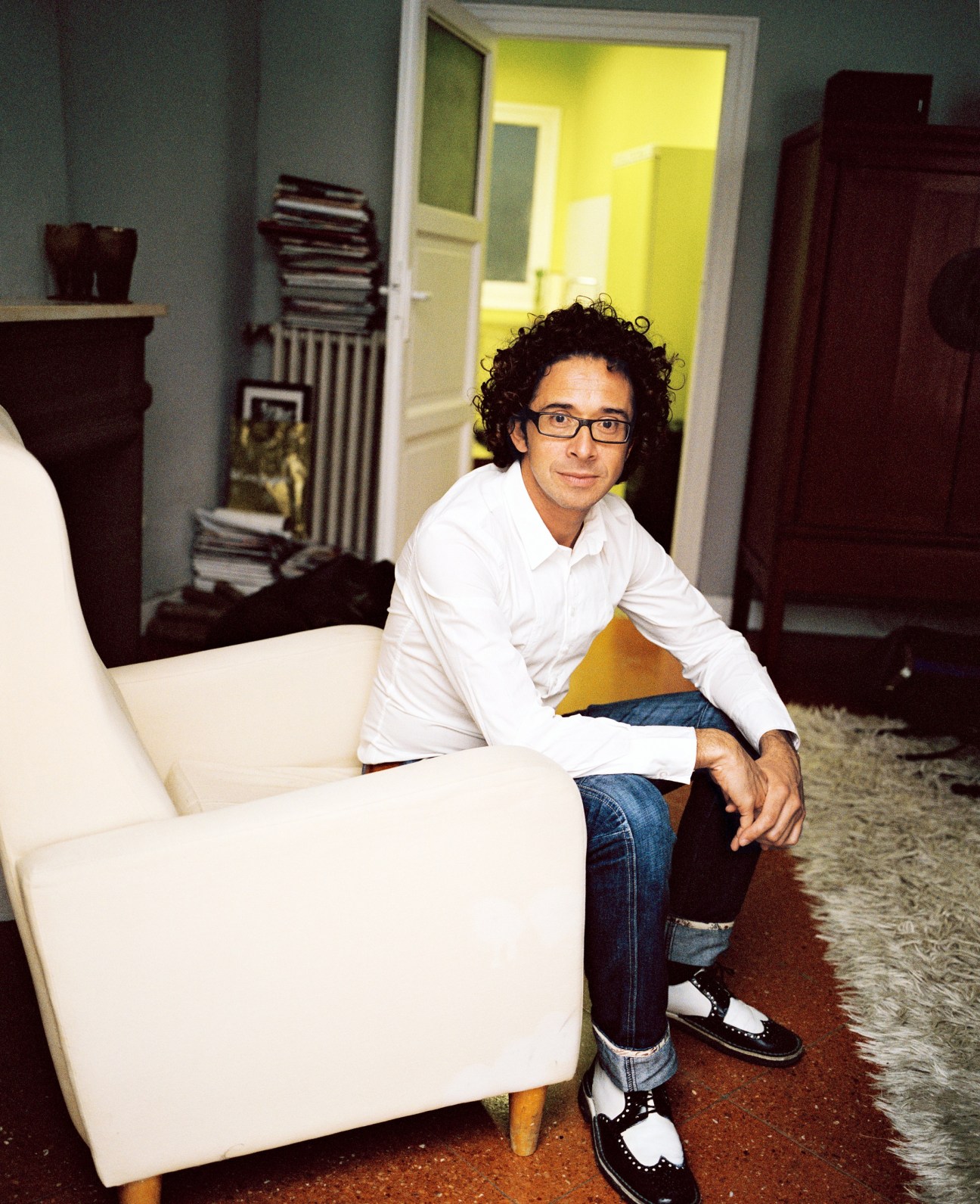 Ramón Barrera, a restaurateur, sitting on a white chair in a modern apartment interior with yellow-painted walls.
