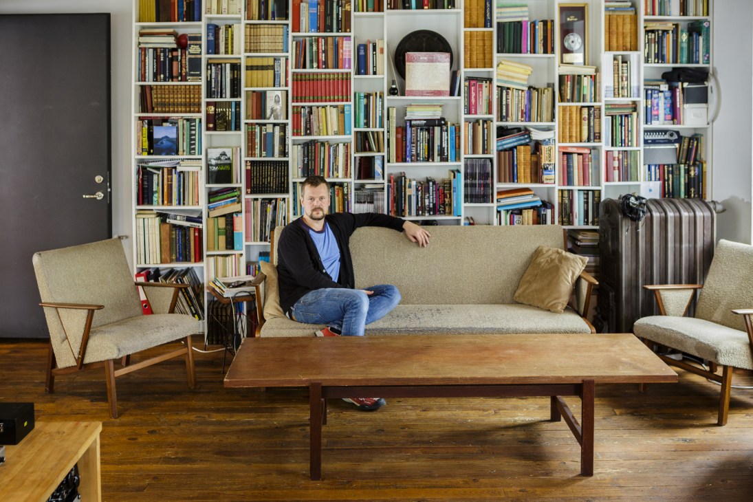 Birgir Jón Birgisson sits on a beige sofa in front of a floor-to-ceiling bookshelf filled with books and records.