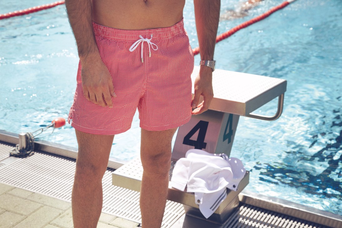 Man in red gingham swim trunks standing at swimming pool edge with lane dividers.