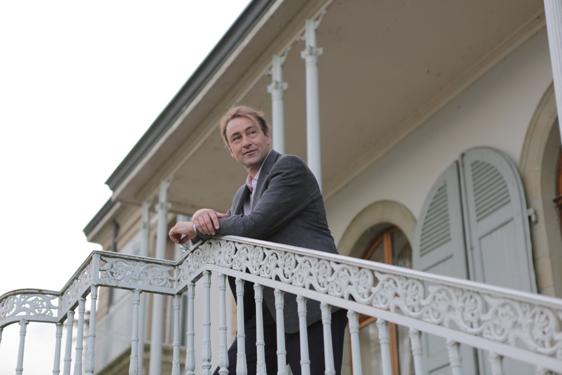 David Harland stands on a balcony of a classical building with white ornamental railings.