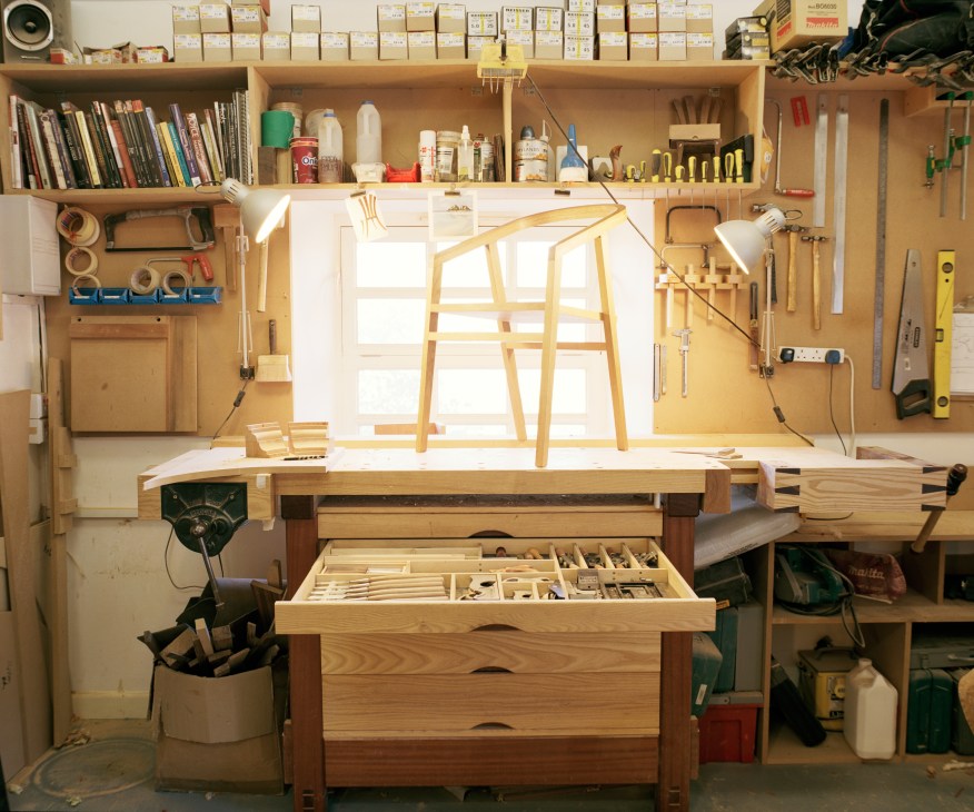 Organized woodworking workshop with handcrafted wooden chair frames on workbench and tools on walls.