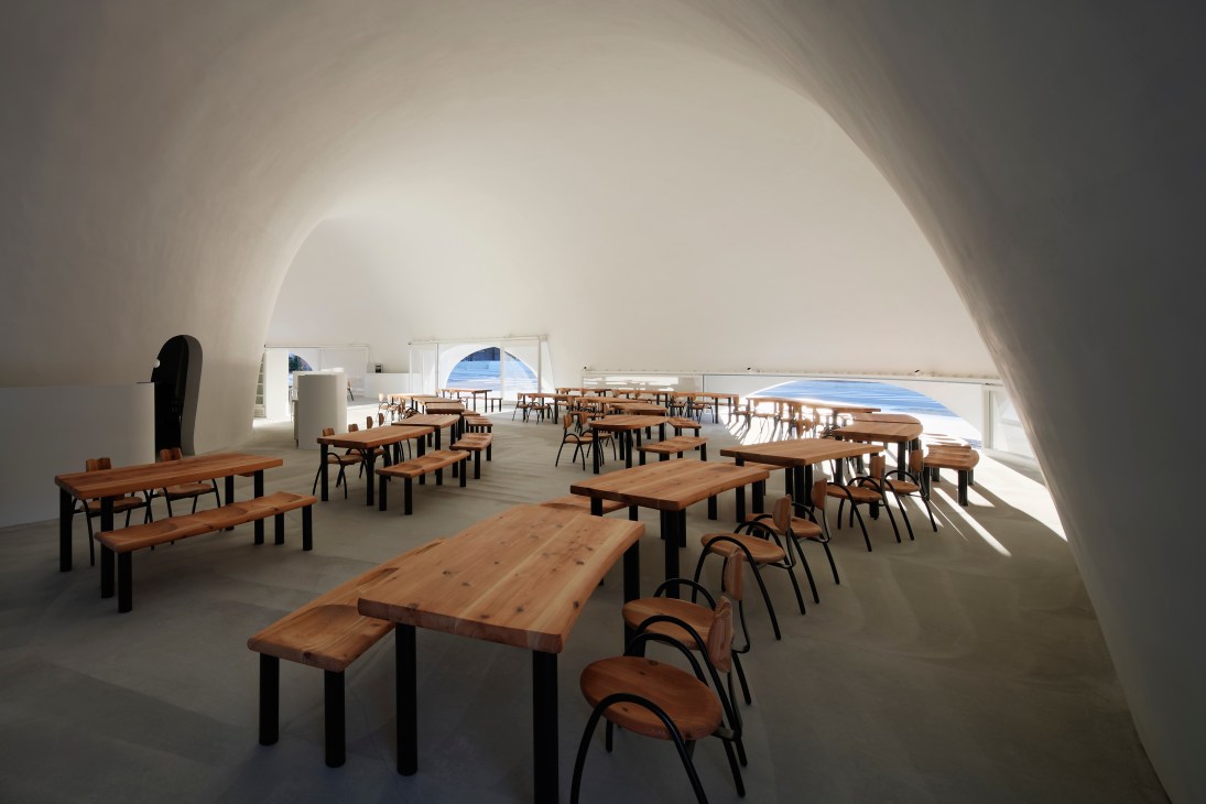 Interior of Hoto Fudo restaurant with white curved architecture, wooden tables and chairs, and large windows overlooking blue water.