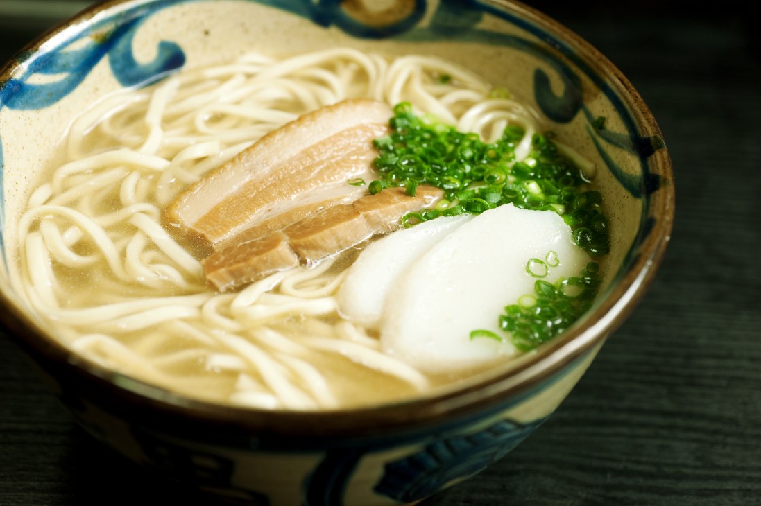 Bowl of Okinawa soba with noodles, broth, pork, egg and green onions in decorative ceramic bowl.