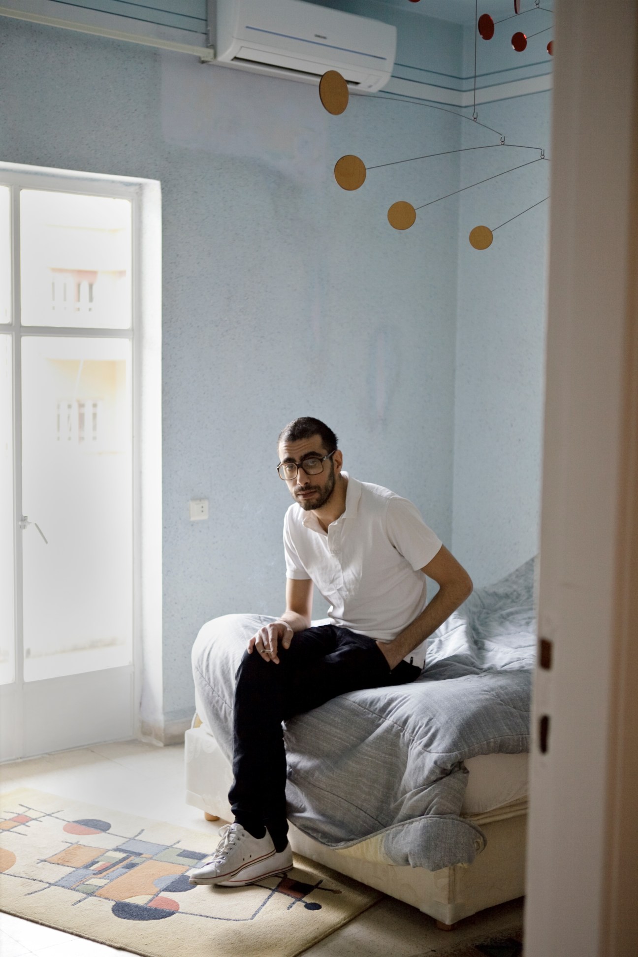 Musician Charbel Haber sits on a bed in a minimalist bedroom with light blue walls and hanging bronze mobile decorations.