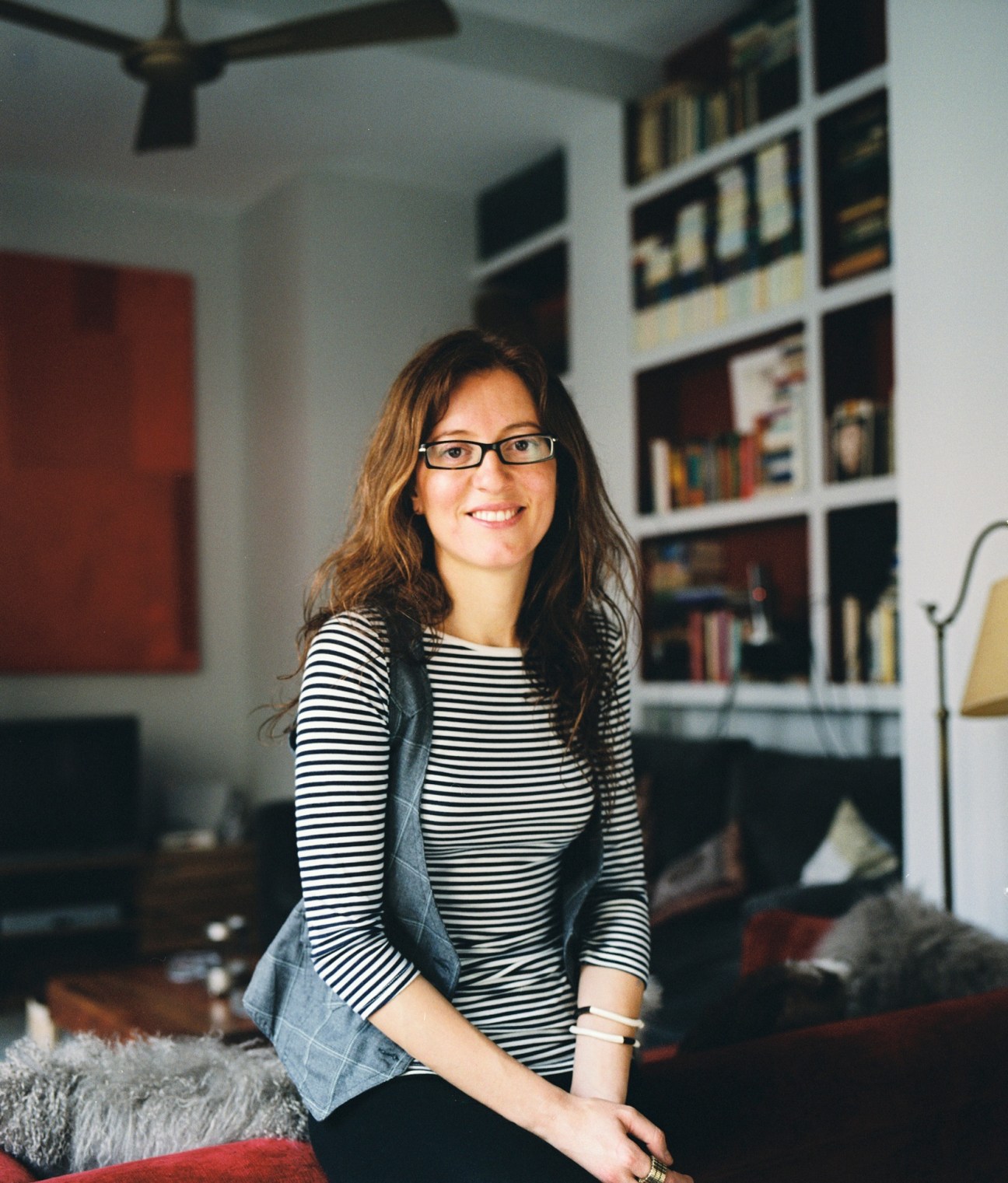 Woman with glasses and long brown hair smiling in a modern living room with shelving and red artwork.
