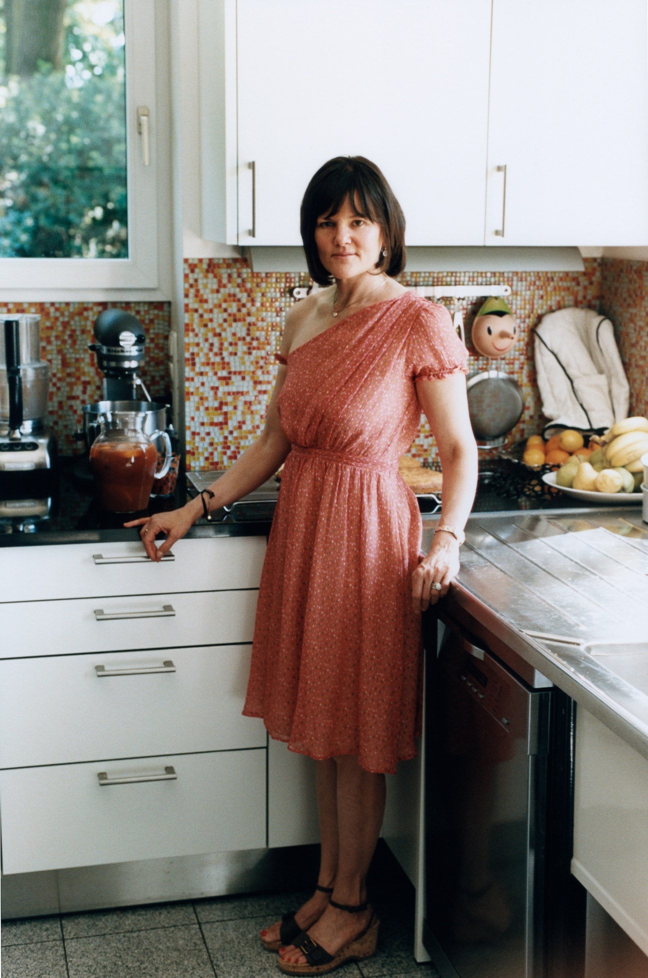 Cynthia Barcomi Friedman stands in her kitchen wearing a rust-colored dress, with colorful tile backsplash and white cabinetry visible behind her.