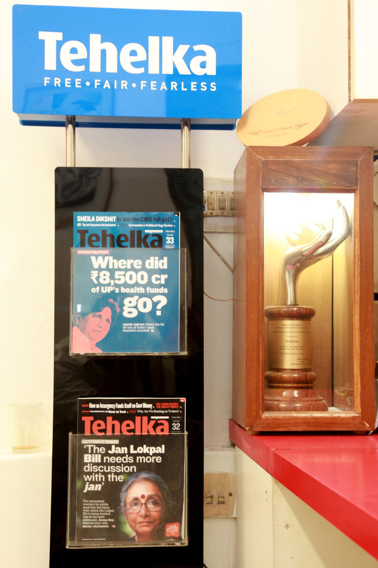 Tehelka magazine covers displayed in a black wall-mounted rack with the blue 'Tehelka Free Fair Fearless' logo above.
