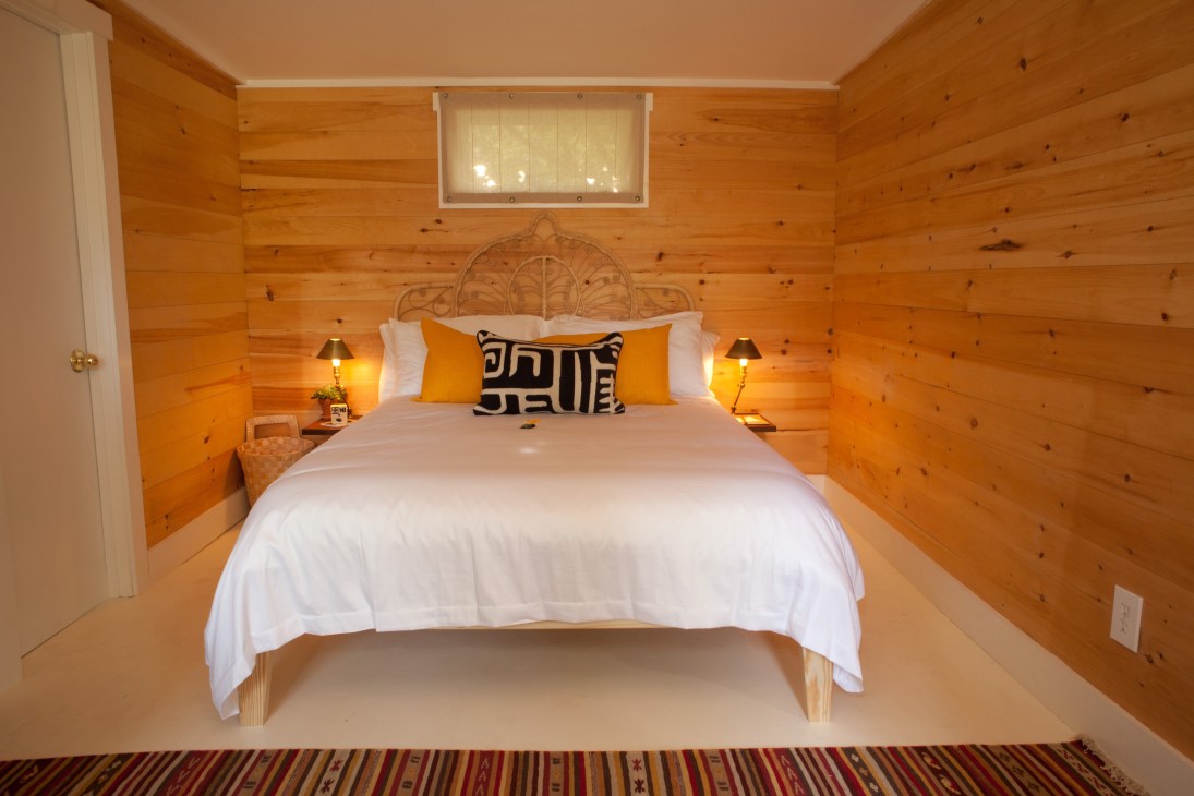 Interior of a wooden cabin bedroom with a bed, yellow pillows, bedside lamps and a window.