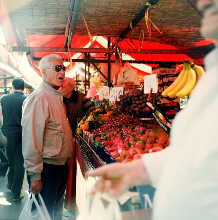 An elderly man in sunglasses and pink shirt browses fresh produce at Porta Palazzo market in Turin.
