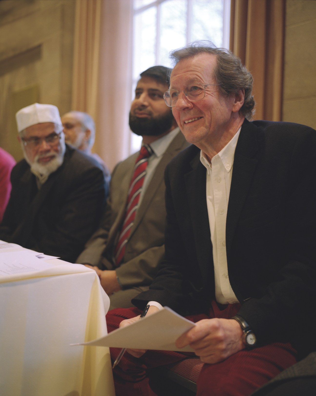George Ferguson, an older man with glasses wearing a dark blazer and white shirt, smiles while seated at a table with other people in formal attire in the background.