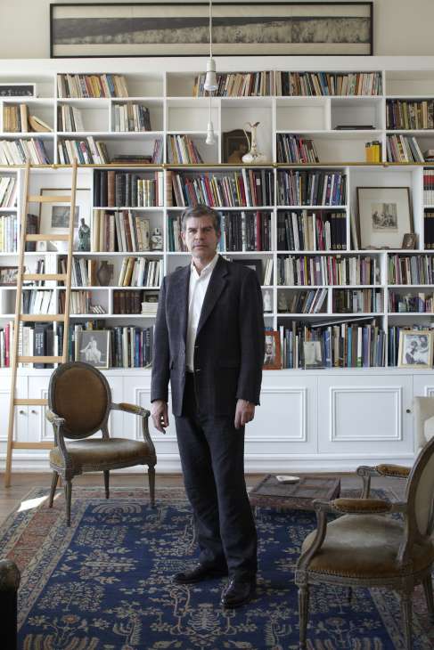 Emilio Lamarca stands in gallery space Casa E before bookshelves, wearing a dark blazer and white shirt.