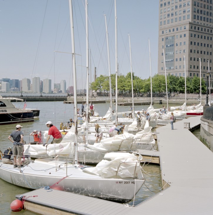 Sailboats docked at Battery Park City jetties with Manhattan skyline and green trees in background.