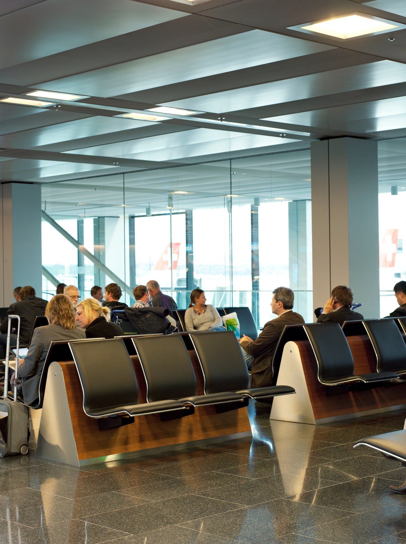 Modern airport lounge with passengers seated in dark leather chairs beneath bright ceiling panels and large windows overlooking runways.