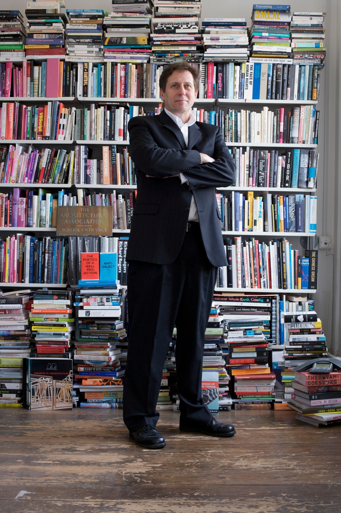 Brett Steele, Director of the Architectural Association School of Architecture, stands in his London office before a floor-to-ceiling bookshelf filled with colorful design and architecture books.