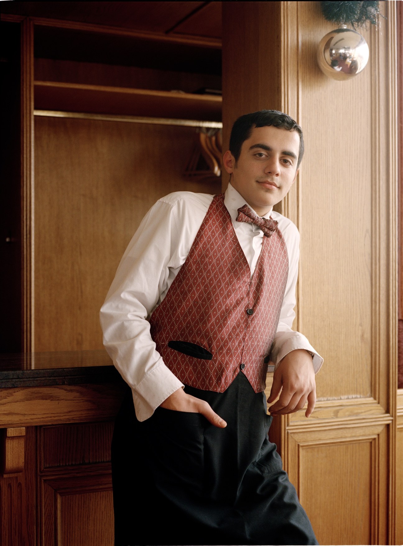 Waiter in formal attire with red patterned vest and bow tie standing in an elegant wood-paneled interior