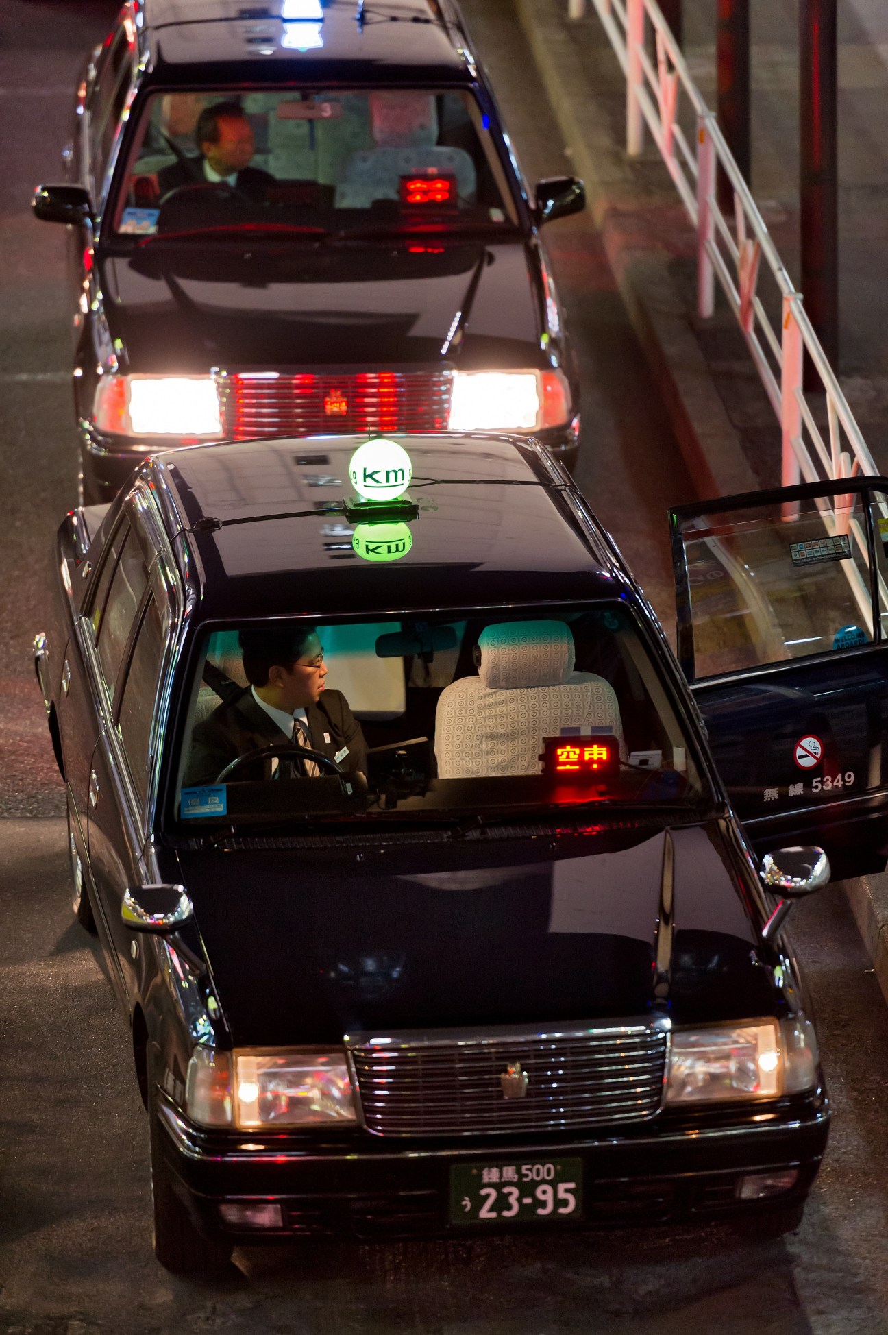Tokyo taxi drivers in black sedans queue on a nighttime street, with illuminated meters and available lights visible.