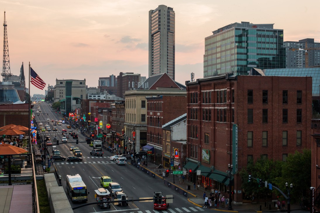 Downtown Nashville cityscape showing Broadway street with historic brick buildings, modern high-rises, and American flag at dusk.