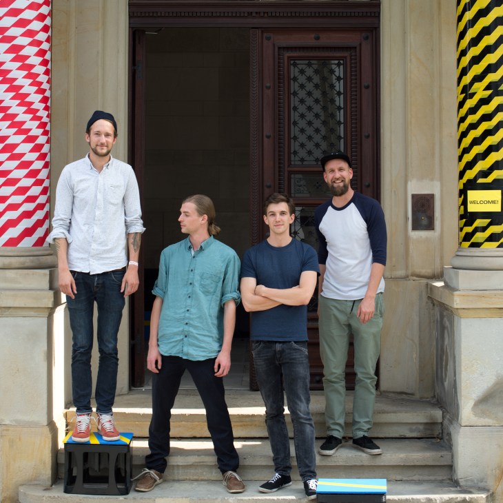 Four men standing in front of a classical building entrance with colorful geometric banners on either side.