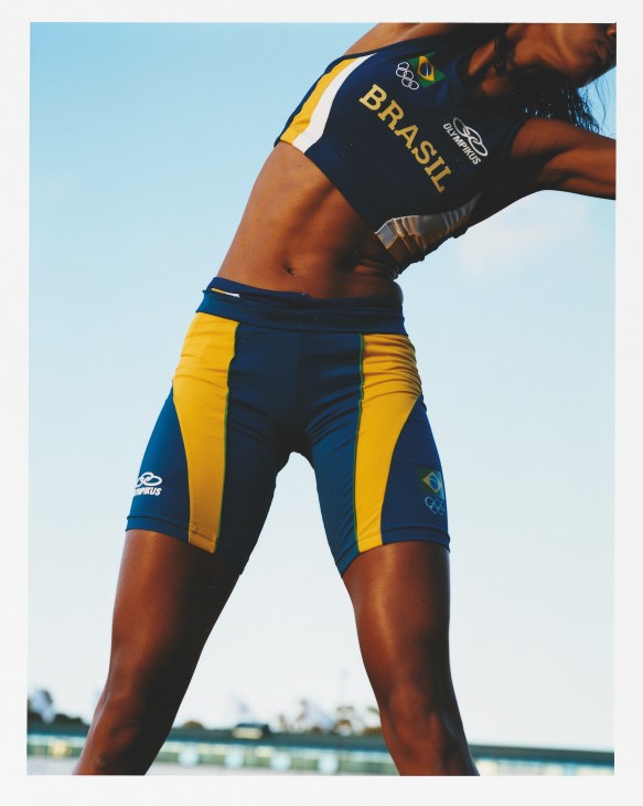 Brazilian athlete in navy and gold tracksuit stretches before a race against clear sky.