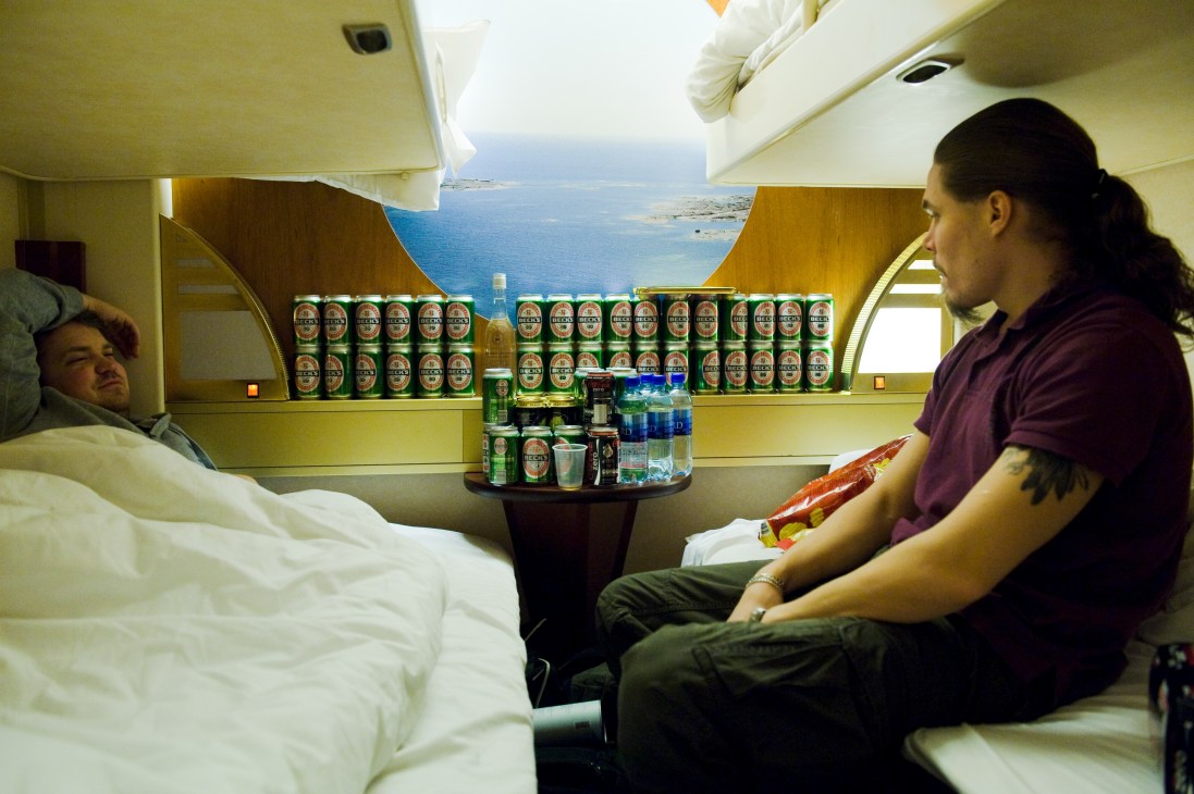 Two men in a ferry cabin stocked with numerous beer cans on a shelf, preparing to disembark with duty-free alcohol.