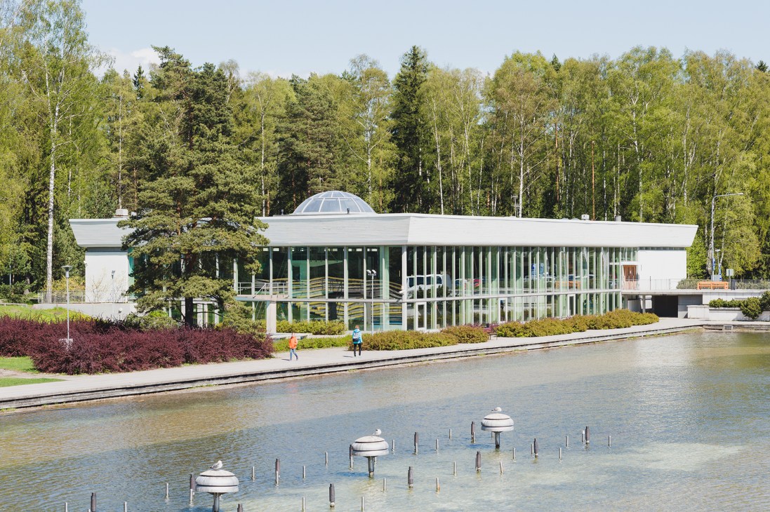 Modern white building with glass walls and dome roof beside a lake with forest backdrop in Tapiola, Finland.