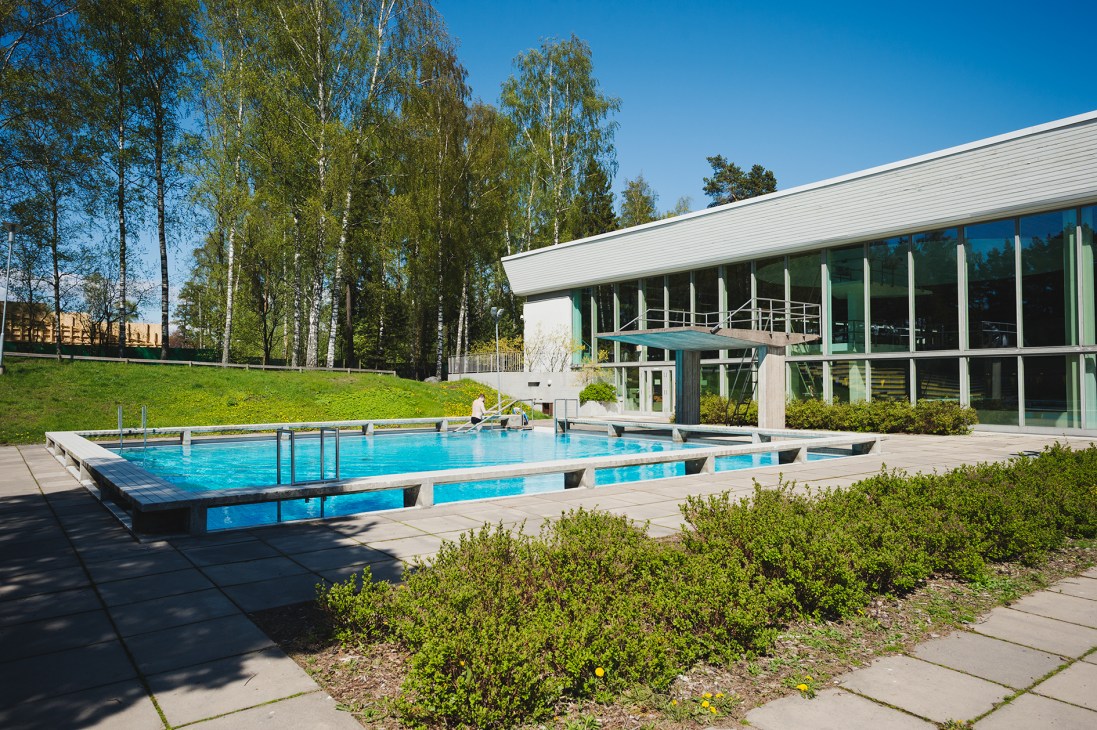 Modern swimming pool with blue water surrounded by green landscaping and contemporary white building in Tapiola, Finland.