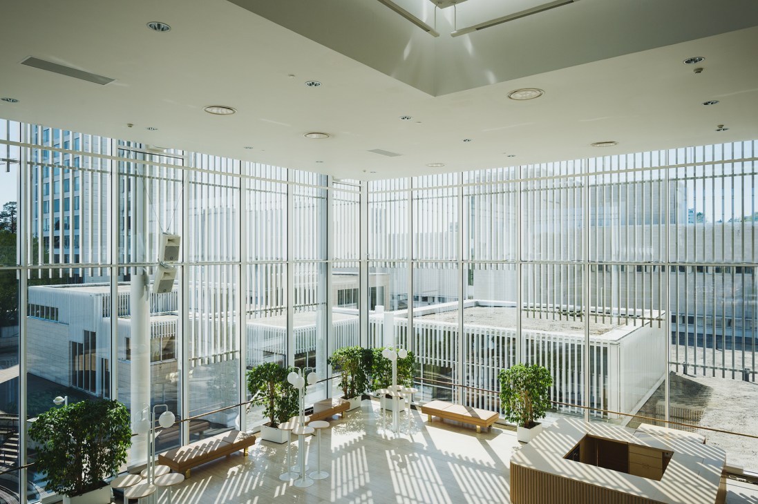 Interior of a minimalist cultural centre with floor-to-ceiling vertical louvers, skylights, potted plants, and wooden seating areas.