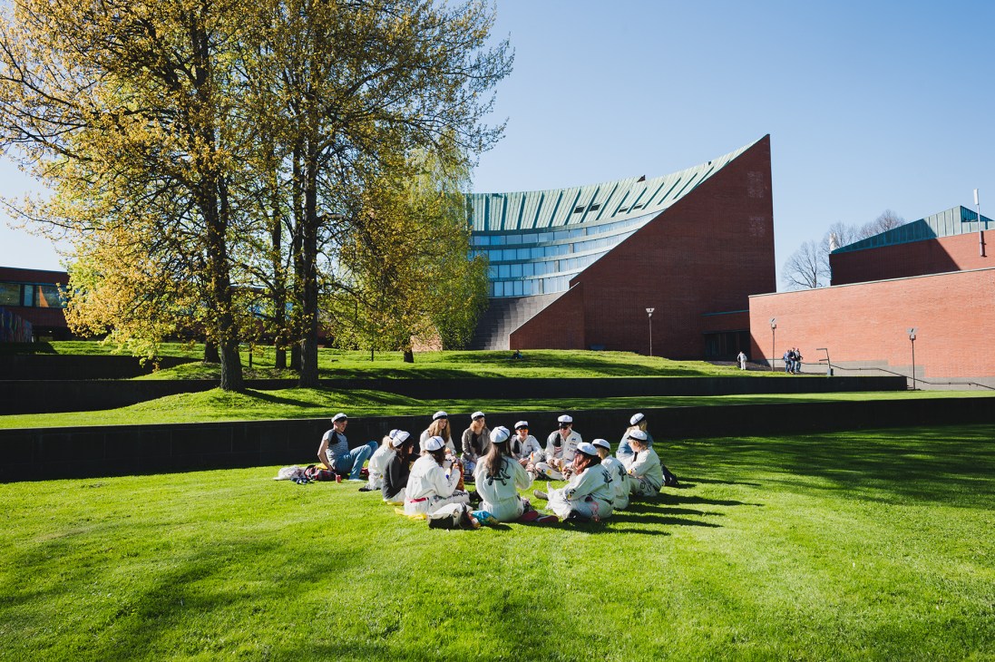Students gathered in a circle on grass outside Aalto University's modernist brick and copper buildings in Otaniemi, Espoo.