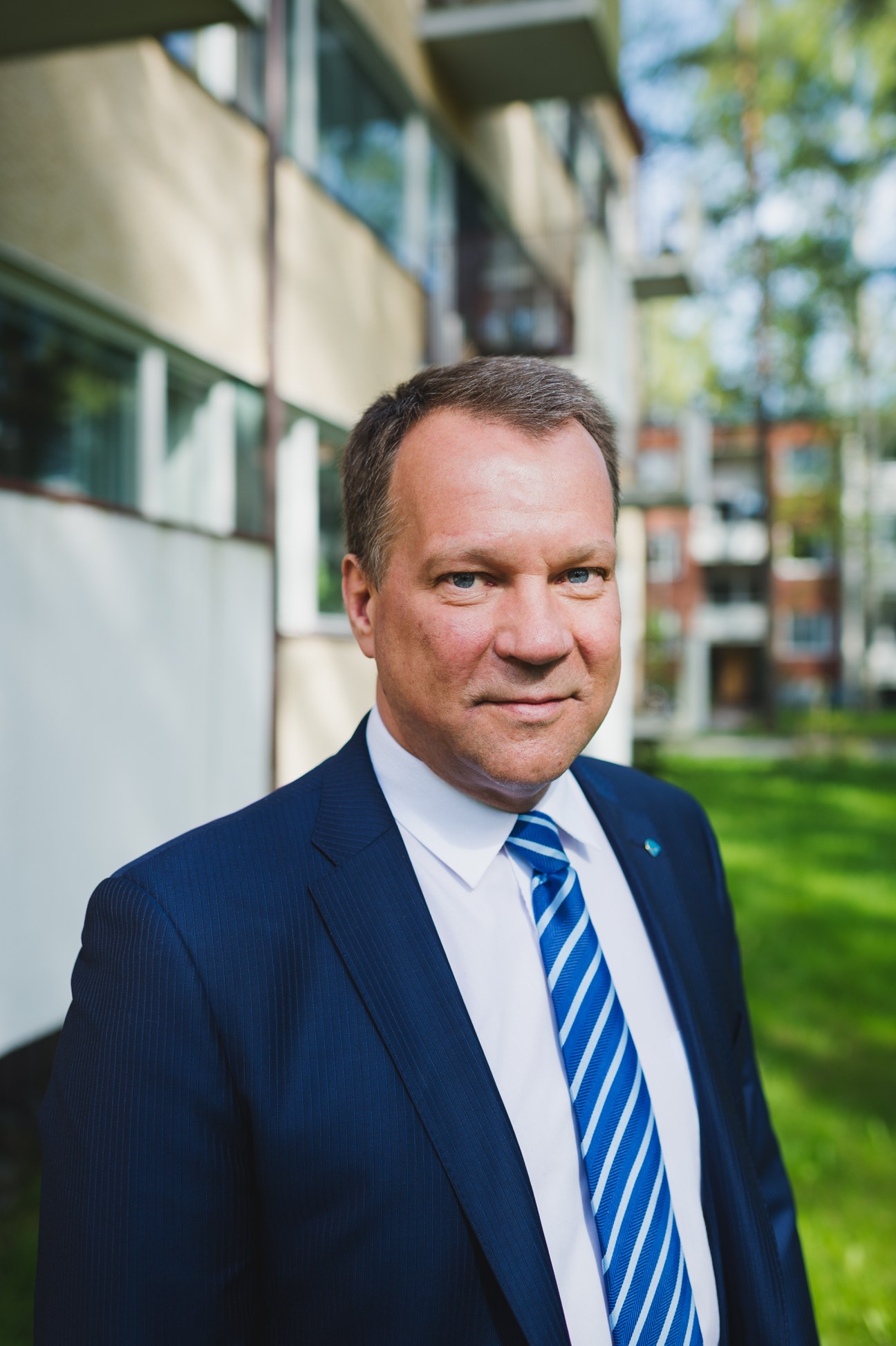 Jukka Mäkelä, mayor of Espoo, standing outside in a dark suit and blue striped tie with modern buildings and greenery in the background.
