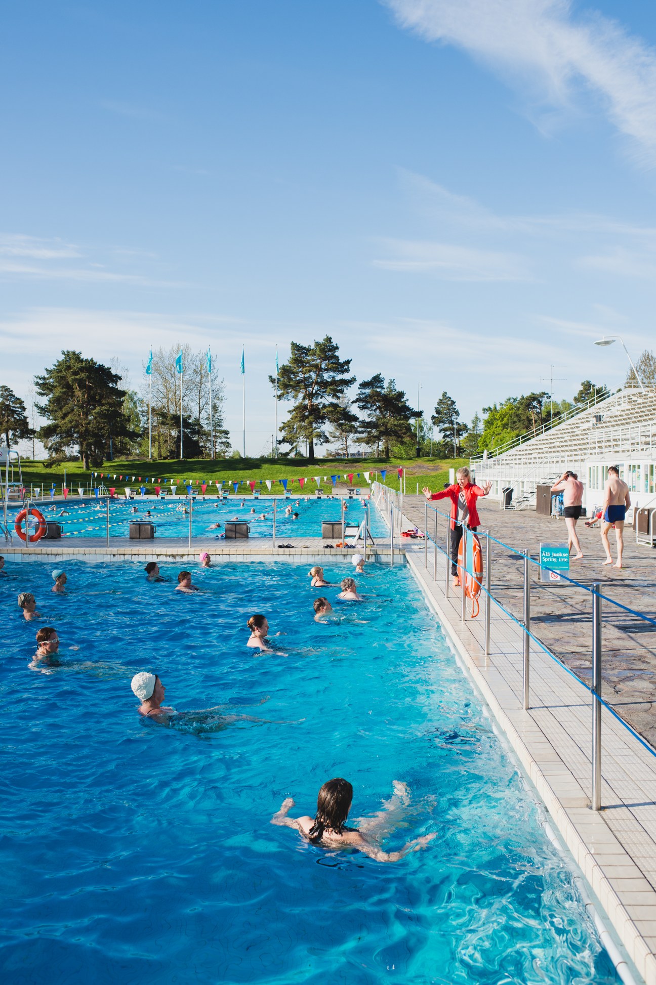 Swimmers exercising in an outdoor pool at Helsinki Swimming Stadium with spectator seating and pine trees visible.