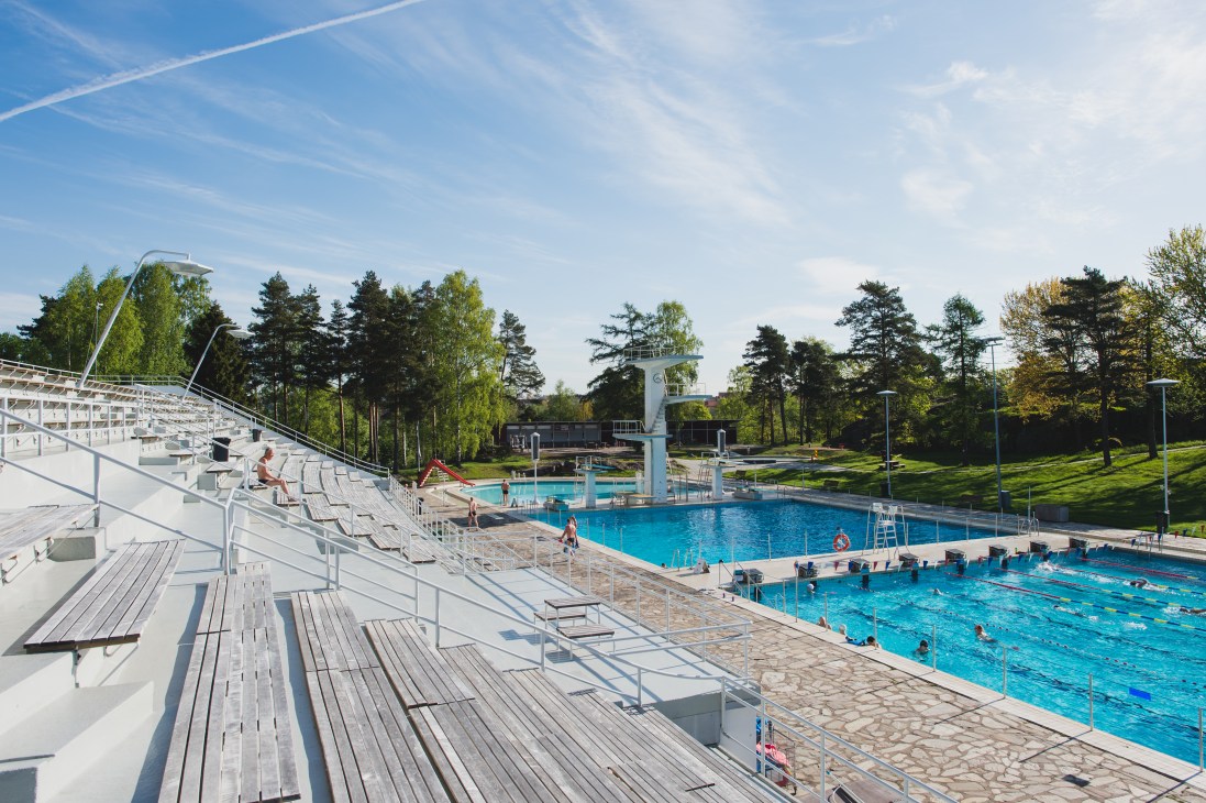 Helsinki Swimming Stadium's outdoor pools with wooden spectator seating, diving tower, and surrounding park trees on a sunny day.