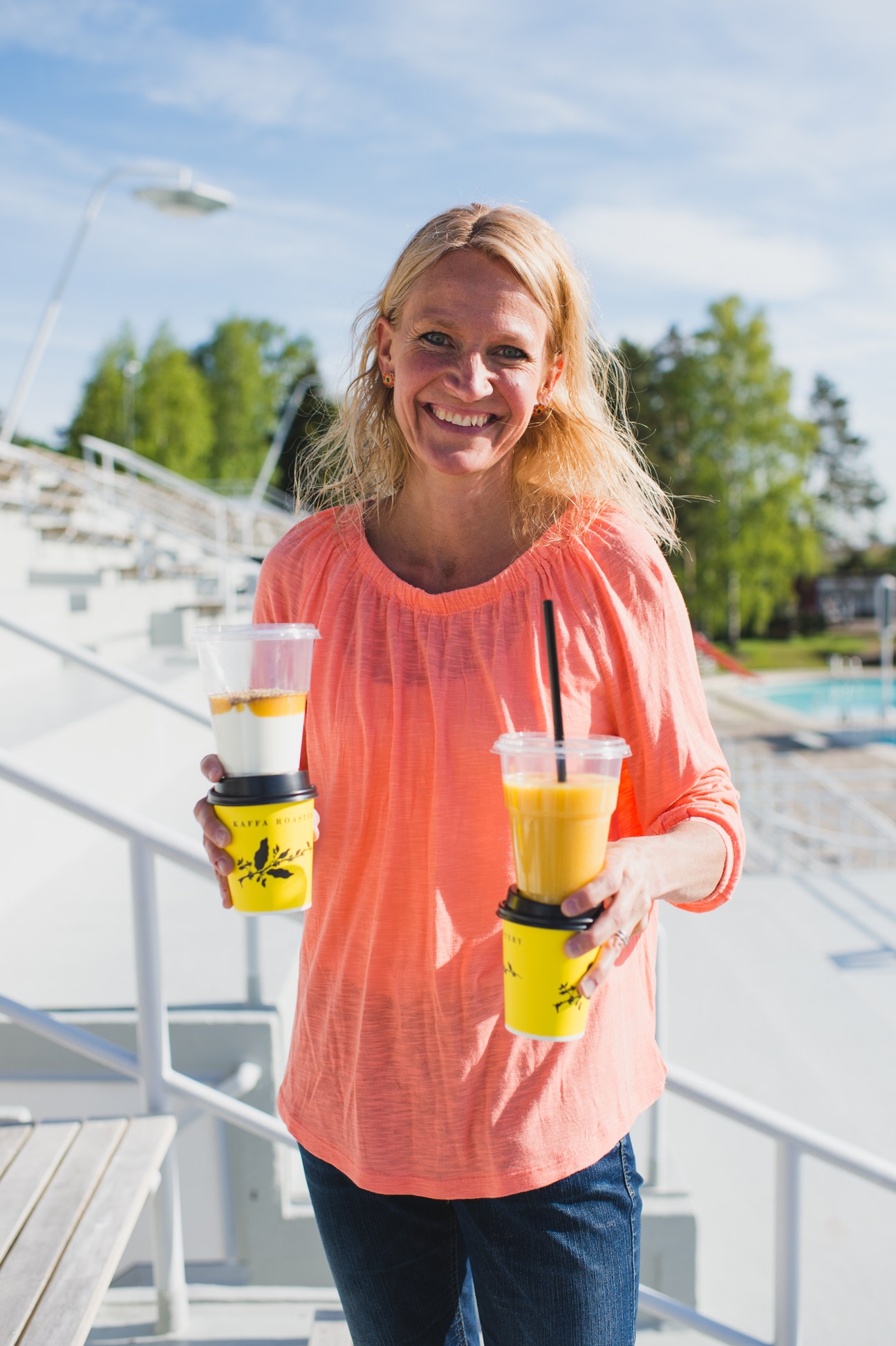 Woman holding drinks at Helsinki Swimming Stadium cafe with pools and trees visible behind her.