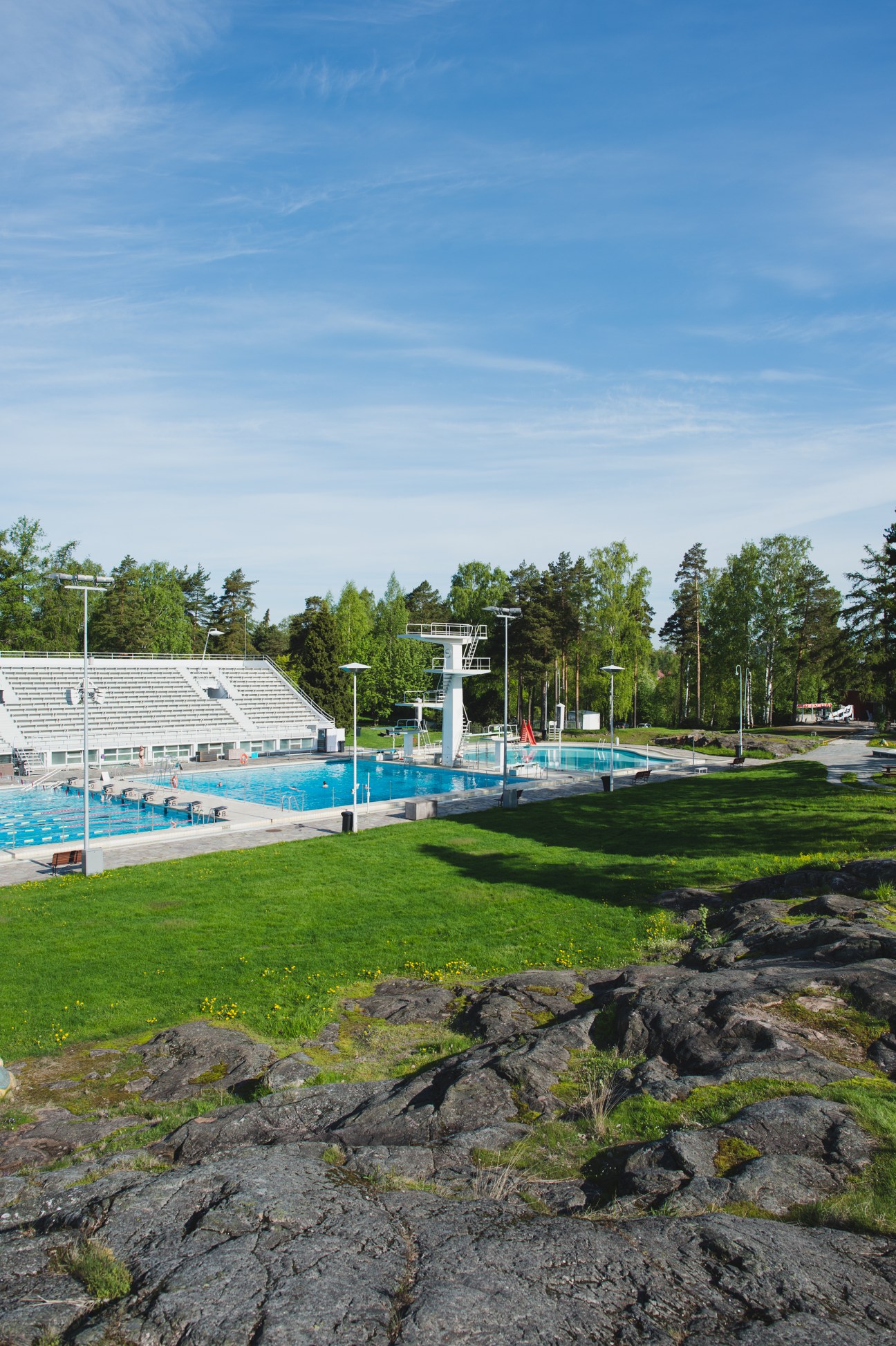 Helsinki Swimming Stadium with blue pools, diving towers, and surrounding parkland with trees and rocky outcrops.