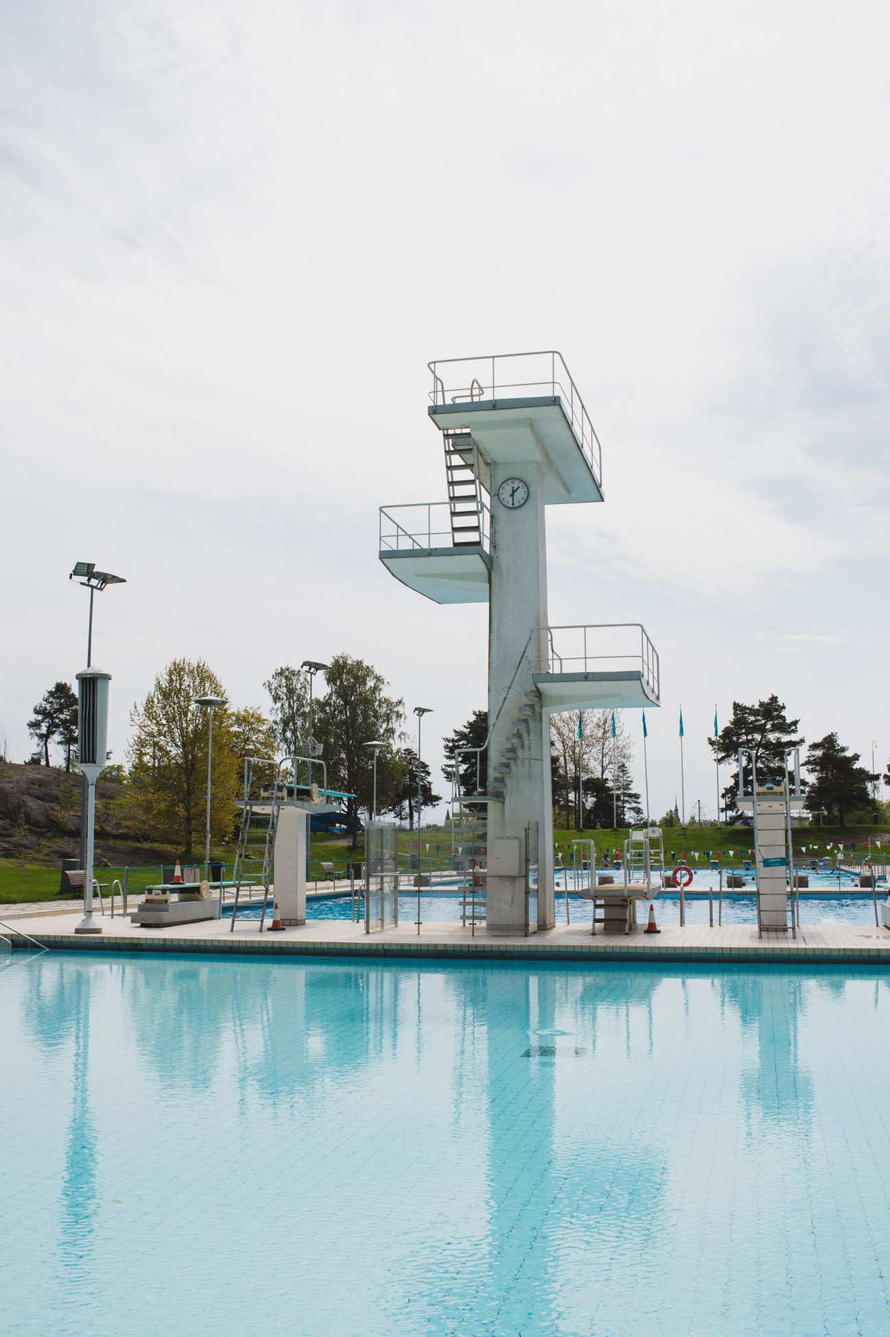 Diving tower with multiple levels overlooking a heated turquoise swimming pool surrounded by green lawns and trees.