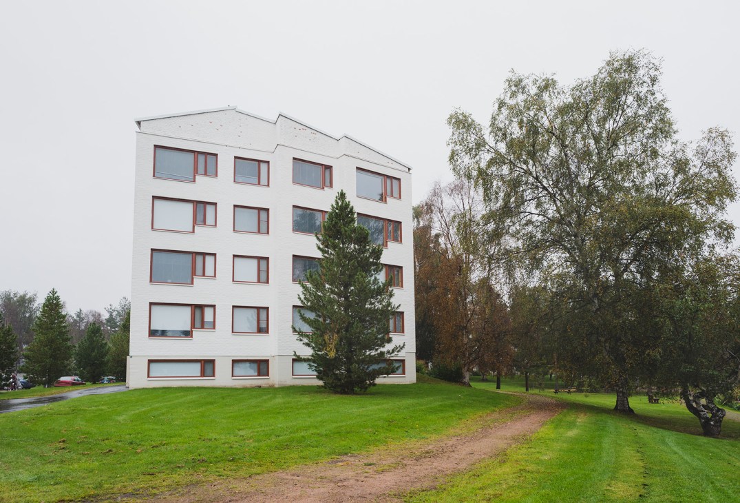 Four-storey white apartment building with red-framed windows set in a green landscape with birch trees.