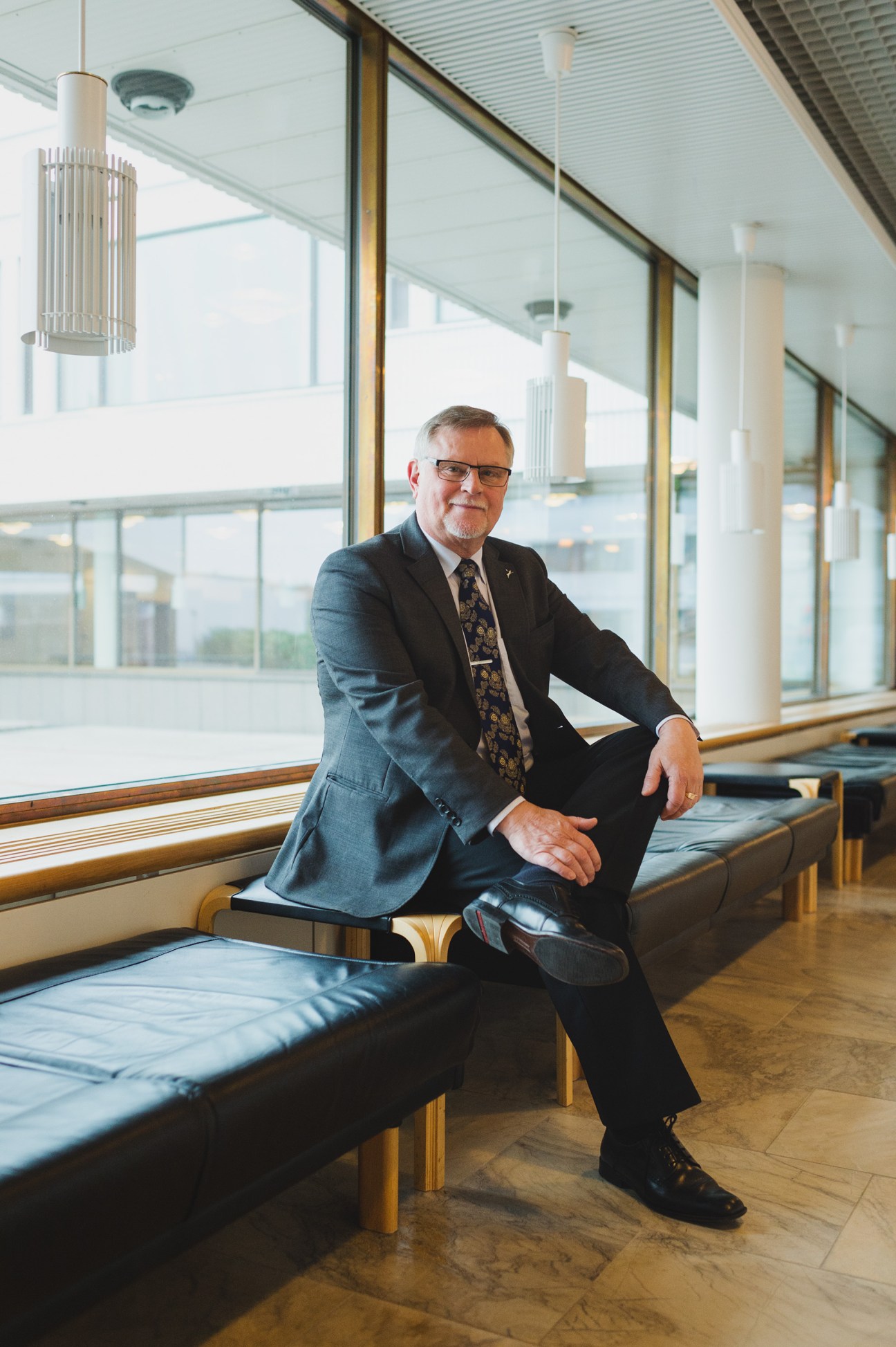 Mayor Esko Lotvonen sits on a bench by large windows in a modernist interior with pendant lighting.