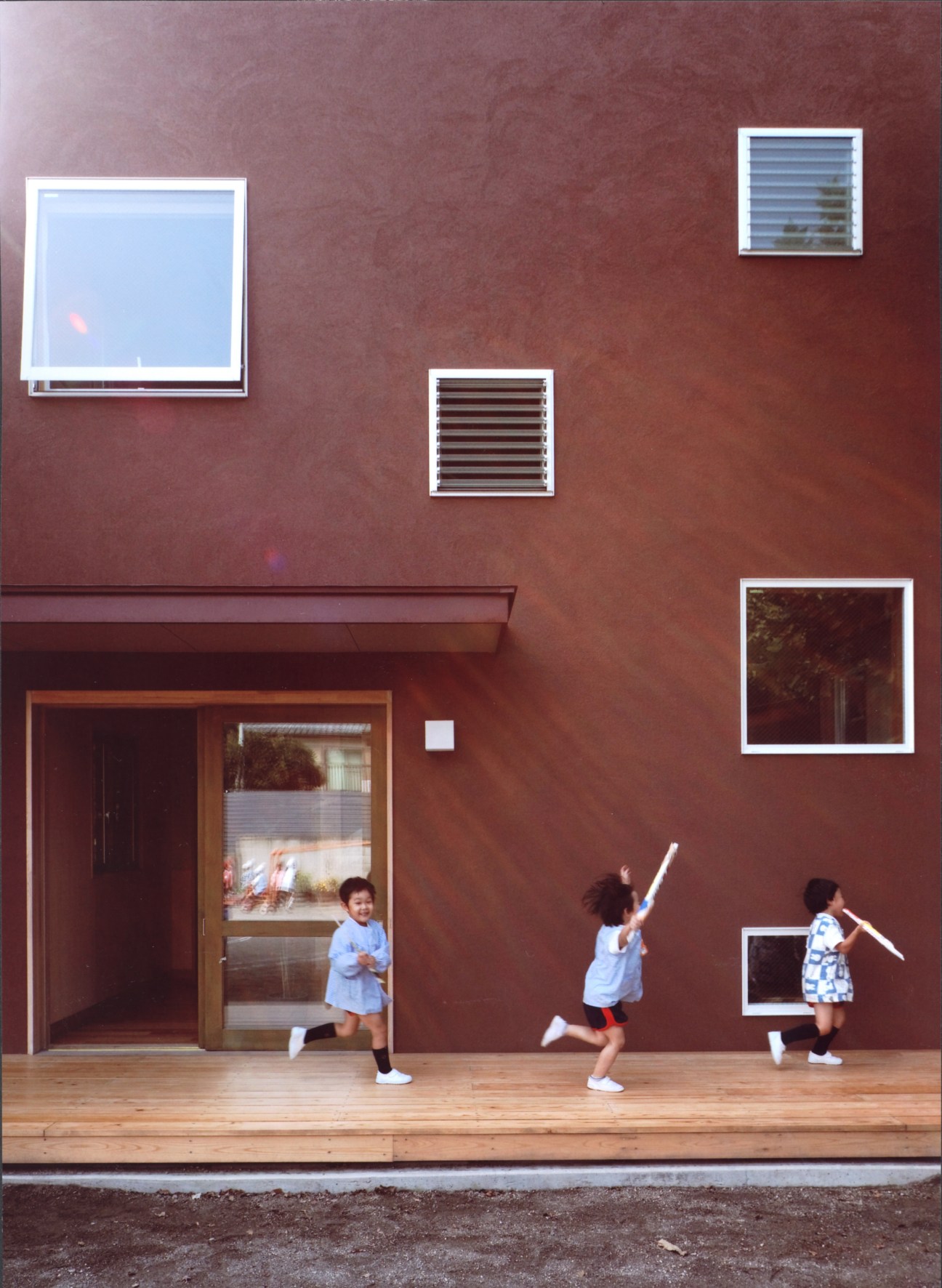 Three young children play on a wooden platform against a terracotta-colored wall with multiple windows at varying heights.
