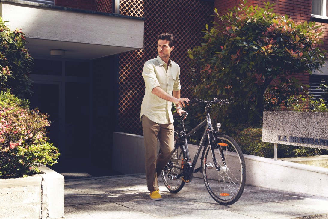 Man in cream shirt and tan trousers with bicycle outside modernist building with brick facade.