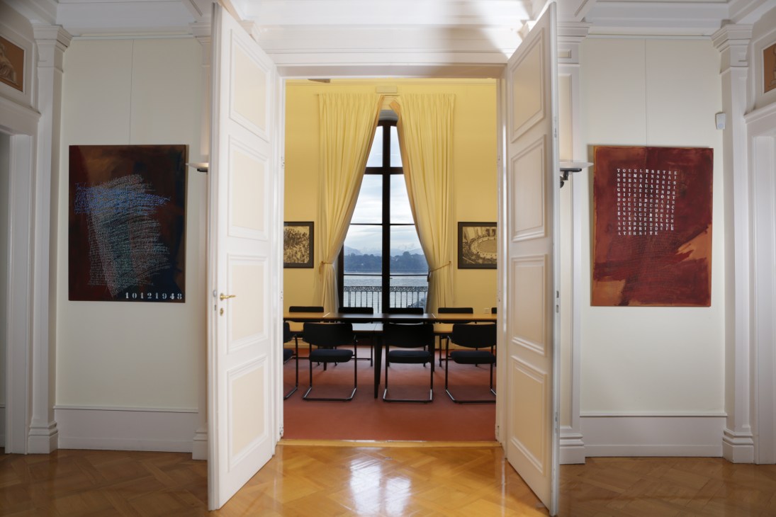 Interior hallway with yellow-walled room featuring large windows overlooking water, modern chairs, and contemporary artwork on walls.
