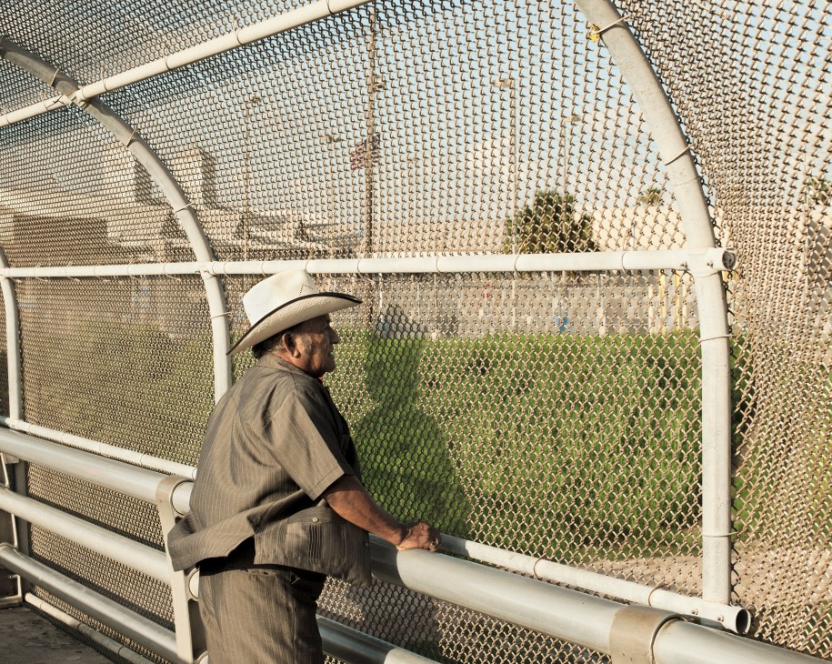 Man in cowboy hat stands at metal border fence on official crossing bridge in Texas.