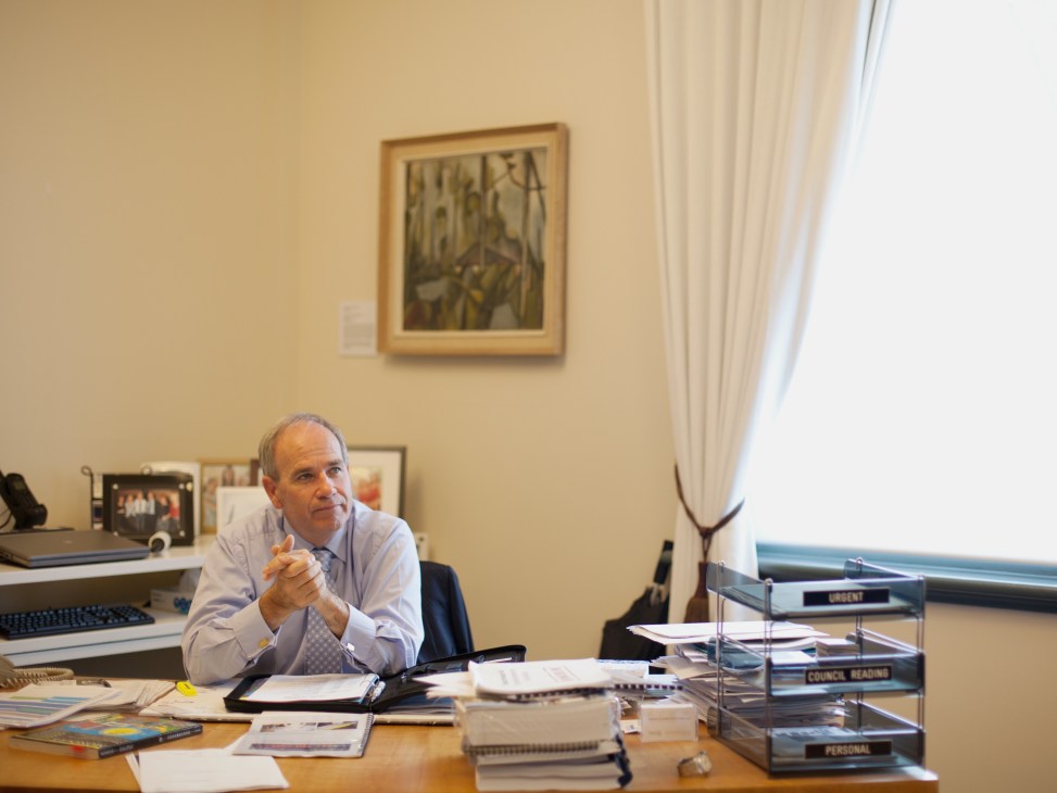 Man in light blue shirt sitting at desk in office with papers and computer.