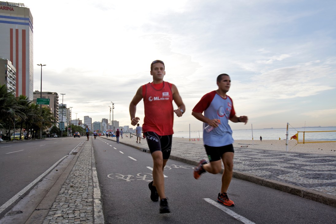 Two men jogging along a beachfront promenade in Rio de Janeiro with city buildings and water visible.