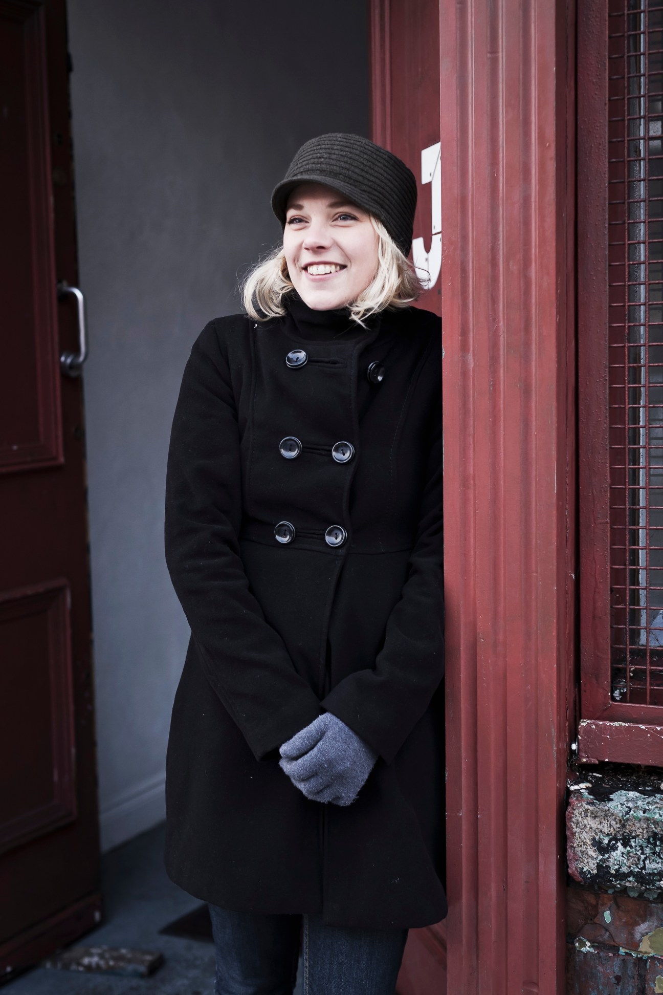 Artist Ella Bertilsson smiles while standing by a red door wearing a black coat and gray hat.