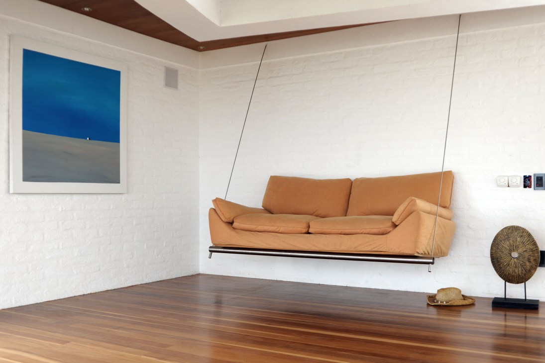 Modern living room with suspended tan leather sofa, wooden floor, and blue abstract artwork.