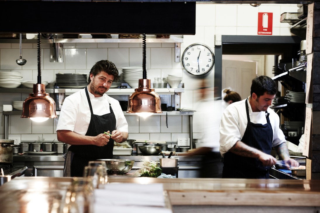 Two chefs work in a modern kitchen at Estelle Bar & Kitchen with copper pendant lights and open shelving.